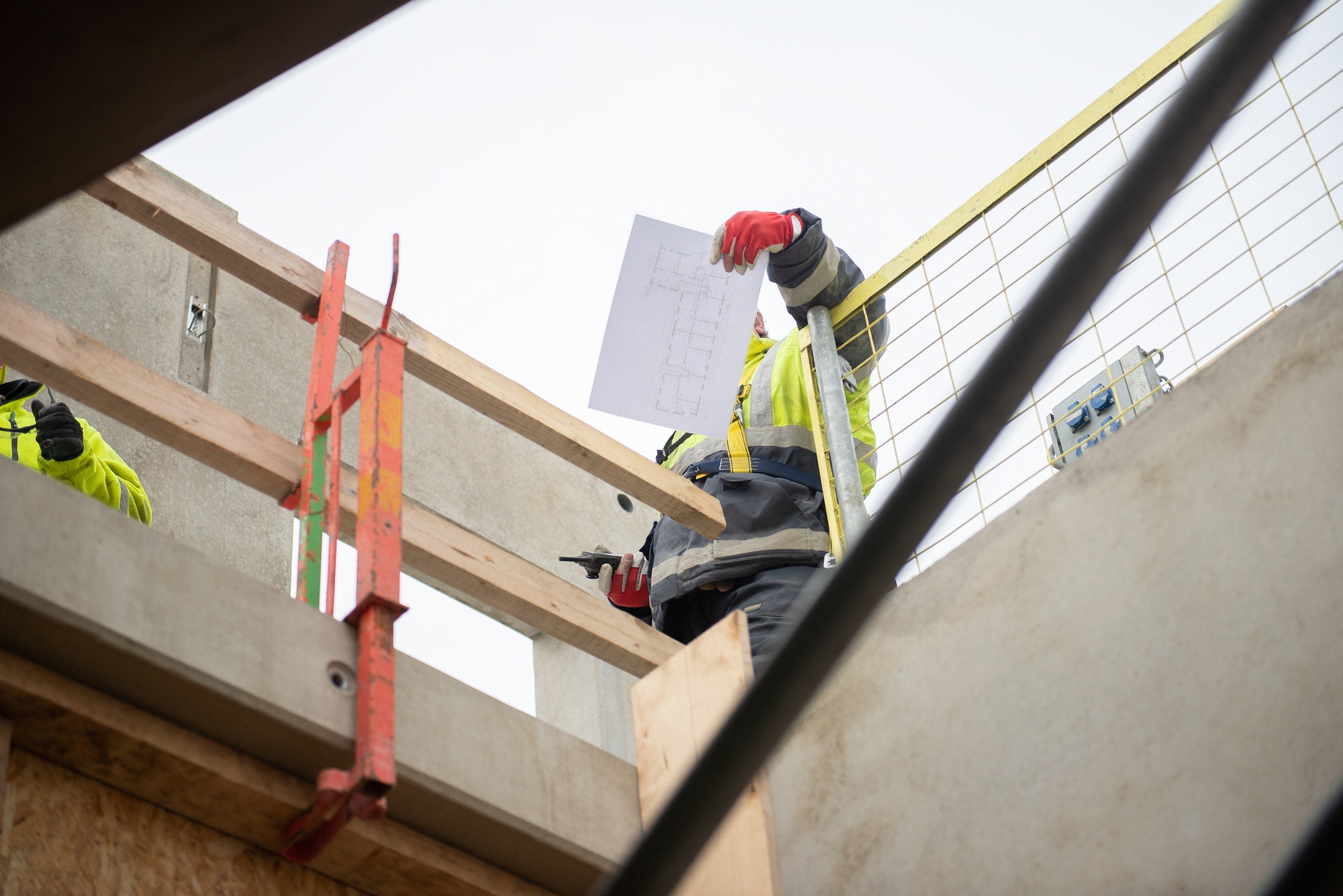 A construction worker with a walkie-talkie and a sheet of design drawings stands on the scaffolding
